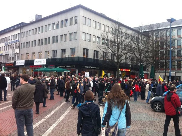 Große Menschenmenge versammelt sich auf einem Platz vor einem mehrstöckigen Gebäude mit Starbucks-Filiale im Erdgeschoss.