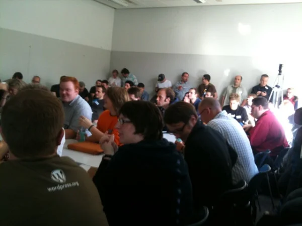 Eine Gruppe von etwa 30 Personen versammelt sich in einem hellen Raum um einen Tisch während einer Veranstaltung oder eines Treffens.
