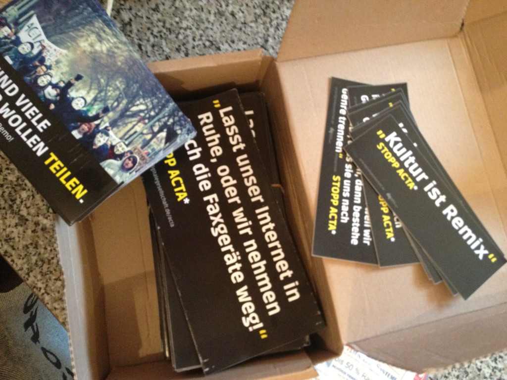 Offene Kartonbox mit schwarzen Flyern und Visitenkarten sowie einer blauen Verpackung auf Betontextur.