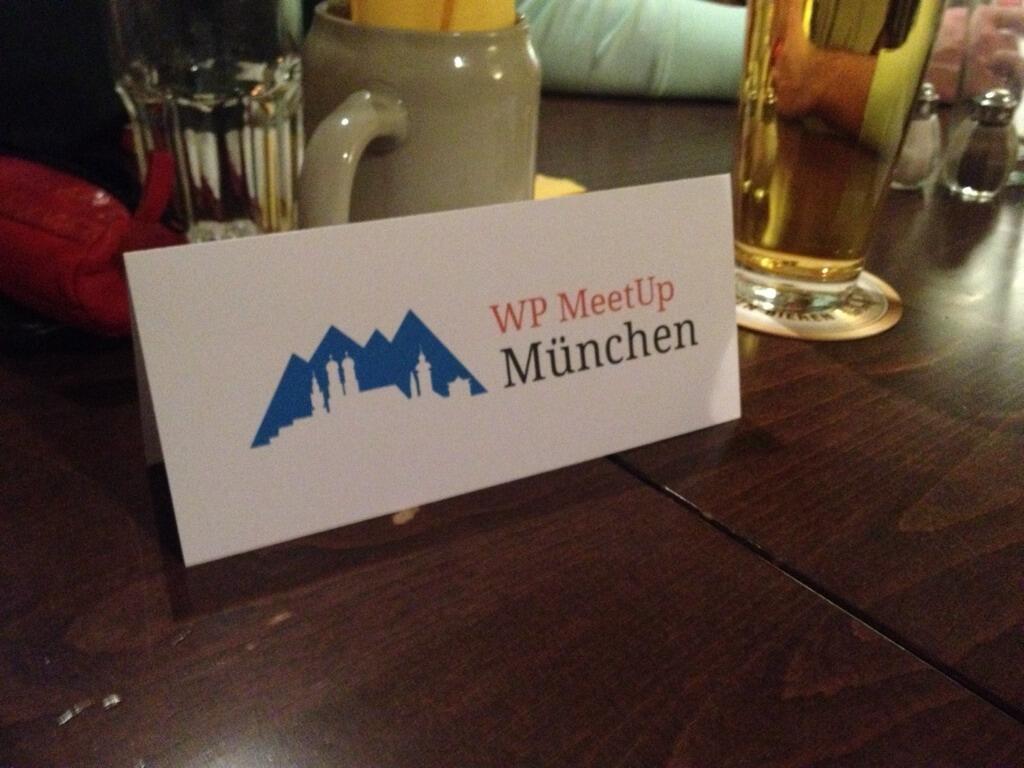 Tischkarte mit dem Logo und Text 'WP MeetUp München' auf einem dunklen Holztisch mit Gläsern und Flaschen im Hintergrund.