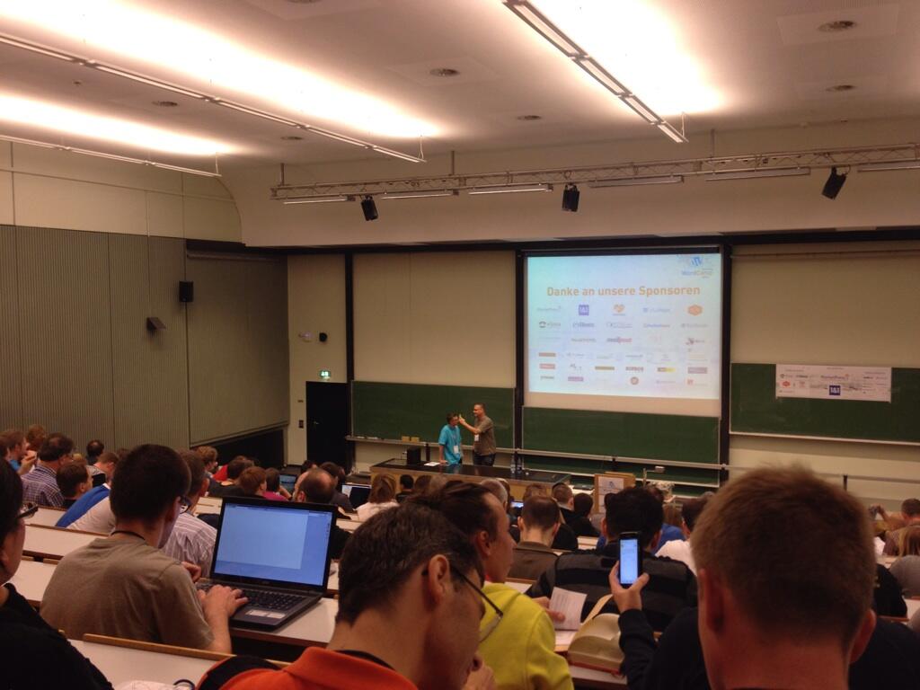 Lecture hall with students seated at desks with laptops, watching two presenters on stage with a projected slide showing sponsor logos and German text 'Danke an unsere Sponsoren'.