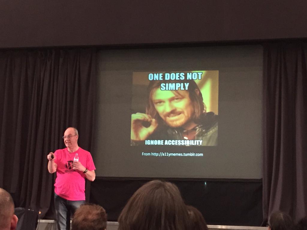 Ein Sprecher in einem pinkfarbenen Hemd präsentiert ein Meme auf einer Leinwand, das die Botschaft "One does not simply ignore accessibility" mit einer bekannten Film-Referenz vermittelt.