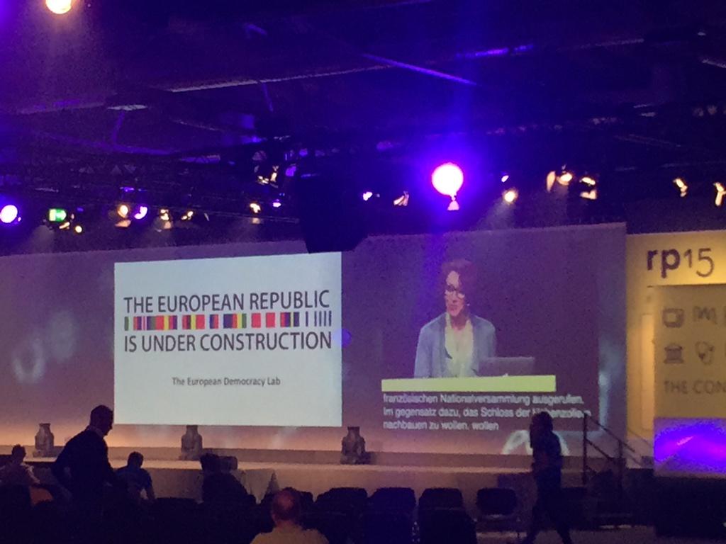 Konferenzpräsentation mit dem Titel 'The European Republic Is Under Construction' vom European Democracy Lab auf einer großen Leinwand in einem Saal mit blauer Beleuchtung.