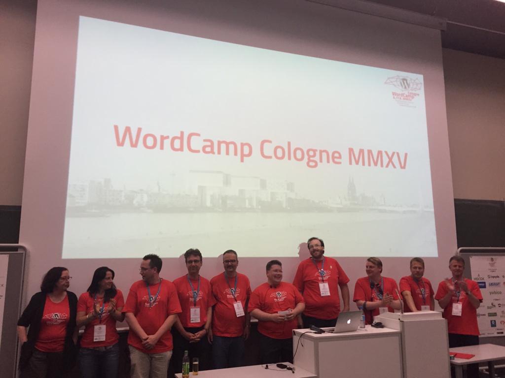 Eine Gruppe von etwa zehn Personen in roten WordCamp-T-Shirts steht vor einer Projektionsfläche, auf der 'WordCamp Cologne MMXV' angezeigt wird.