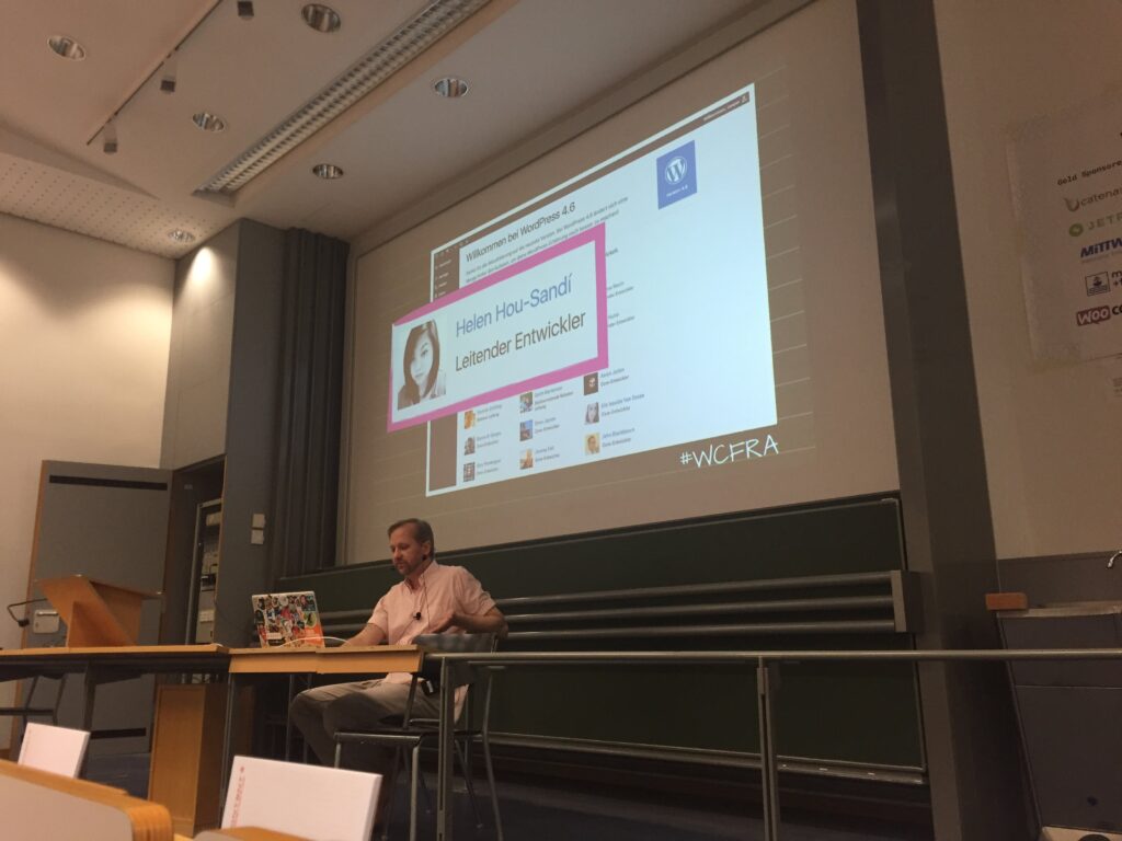 Ein Mann sitzt an einem Tisch vor einer großen Projektionsfläche, die eine WordPress-Präsentation über Helen Hou-Sandi zeigt, mit dem Hashtag #WCFRA sichtbar.