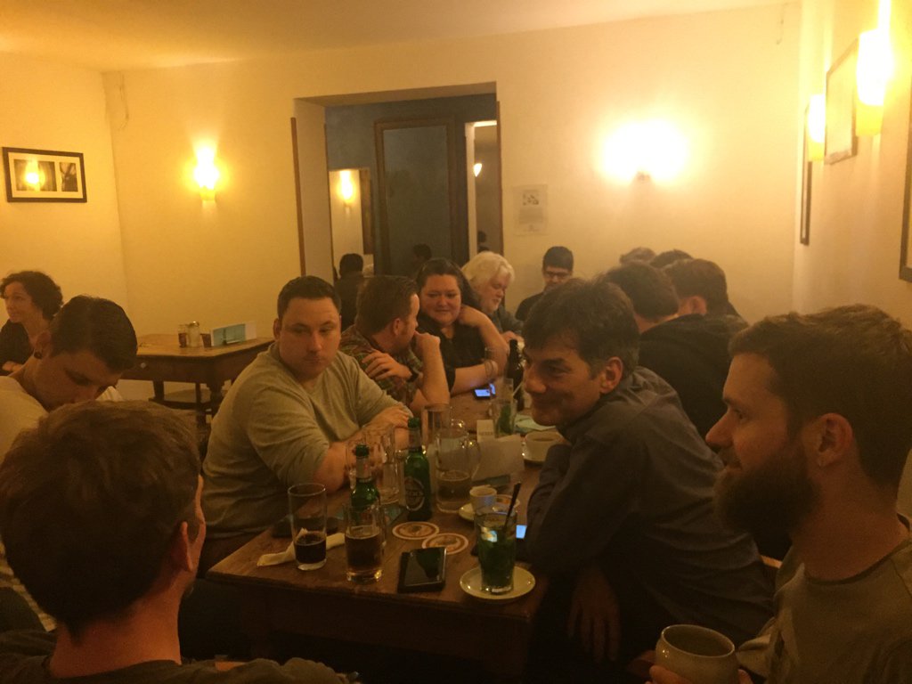 Eine Gruppe von etwa 15 Personen sitzt um einen Tisch in einem warm beleuchteten Raum und genießt gemeinsam Getränke und Gespräche.