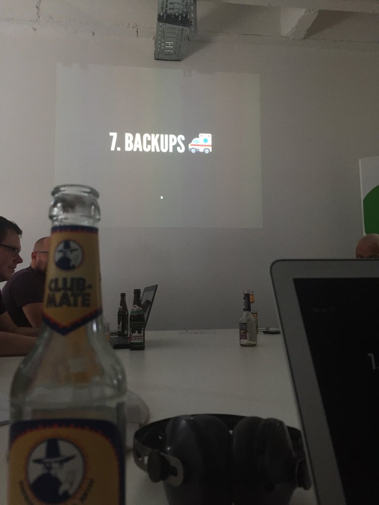 Projektionsbildschirm zeigt "7. BACKUPS" mit einem Lieferwagen-Emoji in einem Konferenzraum, während Personen an einem Tisch sitzen und Bierflaschen vor ihnen stehen.