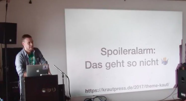 Ein Mann steht neben einem Laptop vor einer Projektionsfläche mit der Aufschrift 'Spoileralarm: Das geht so nicht' und einem Verweis auf eine Webseite.