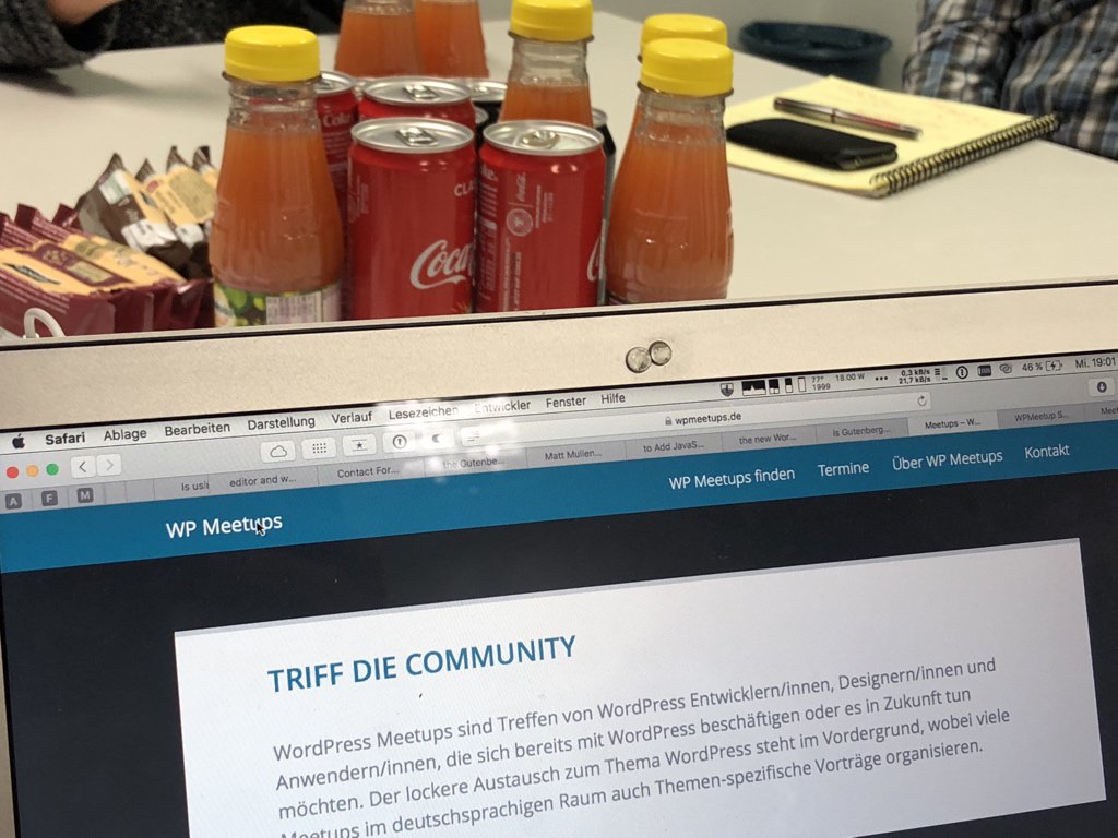 Ein Laptop zeigt eine WordPress Meetups Website mit dem Titel "Triff die Community", während im Hintergrund Getränkeflaschen und Notizblöcke auf einem Tisch stehen.