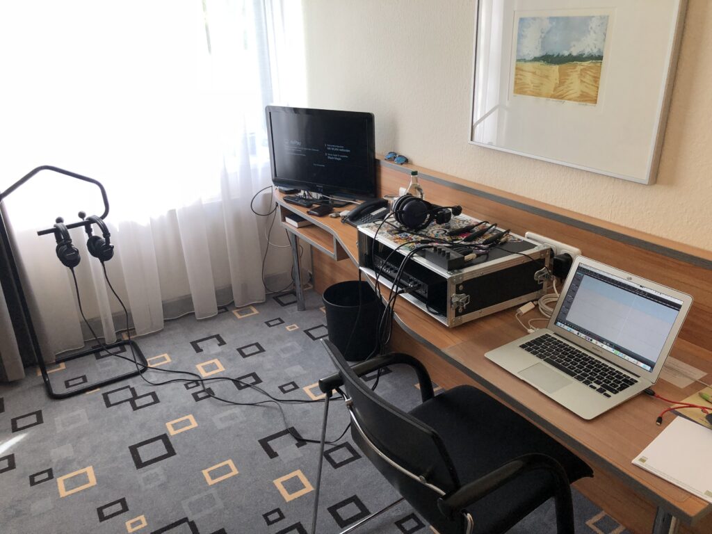 Ein Arbeitszimmer mit einem Schreibtisch aus Holz, auf dem ein Laptop, ein Monitor und professionelle Audio-Ausrüstung stehen, sowie ein Headphone-Ständer und ein schwarzer Stuhl im Vordergrund.