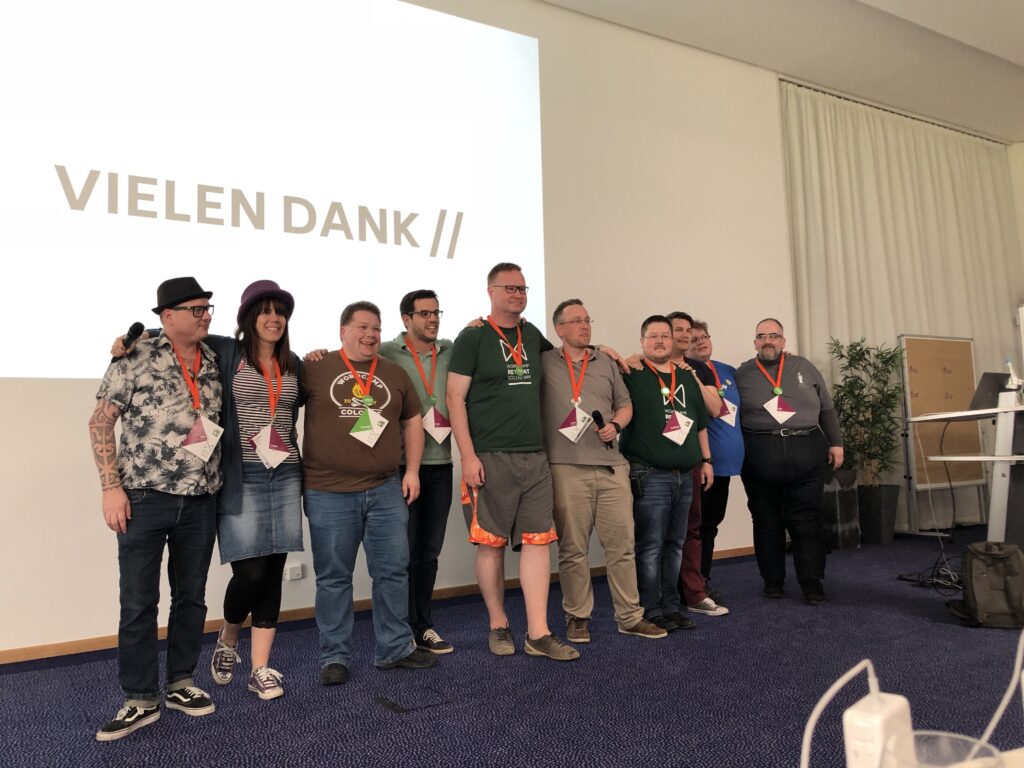 Eine Gruppe von neun Personen mit roten Namensschildern steht zusammen vor einer Projektionsfläche mit der Aufschrift 'VIELEN DANK //'.