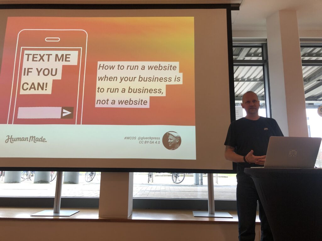 Ein Presenter steht neben einer großen Leinwand mit einer Folie zum Thema Webentwicklung, die den Text "TEXT ME IF YOU CAN" auf einem Telefon-Symbol und "How to run a website when your business is to run a business, not a website" zeigt.