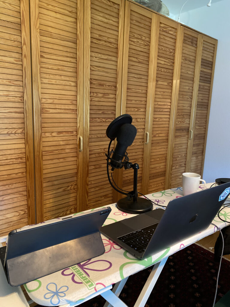 Ein Home-Studio-Setup mit einem Laptop, Tablet, schwarzem Kondensatormikrofon auf Ständer und einer weißen Tasse vor einem hölzernen Raumteiler.