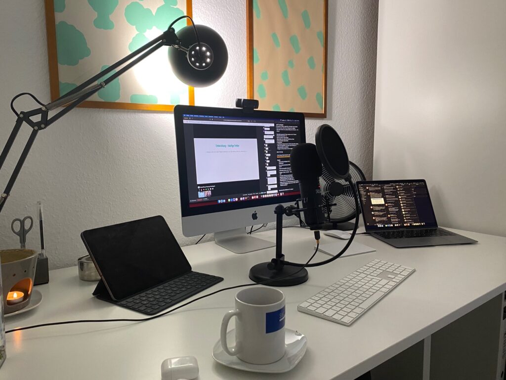 Ein modernes Homeoffice-Setup mit iMac, Laptop, Tablet, Mikrofon und LED-Schreibtischlampe auf einem weißen Schreibtisch.