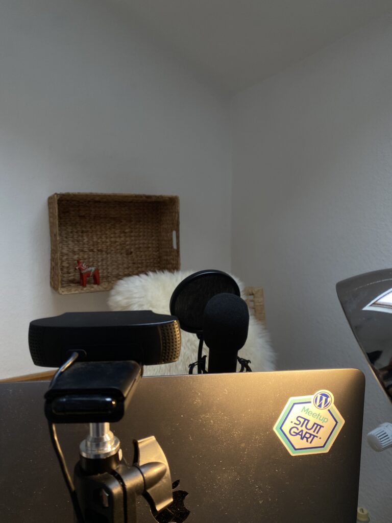 Auf einem Schreibtisch stehen Podcasting-Geräte, darunter ein Mikrofon, Kopfhörer und ein Laptop mit Stuit Cart-Sticker, während im Hintergrund ein geflochtener Wandkorb und ein weißes Fell sichtbar sind.