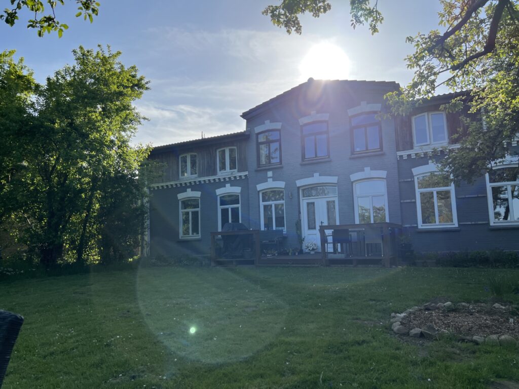 Zweistöckiges Backsteingebäude mit weißen Fensterrahmen und Veranda, umgeben von grünem Rasen und Bäumen bei Sonnenschein.
