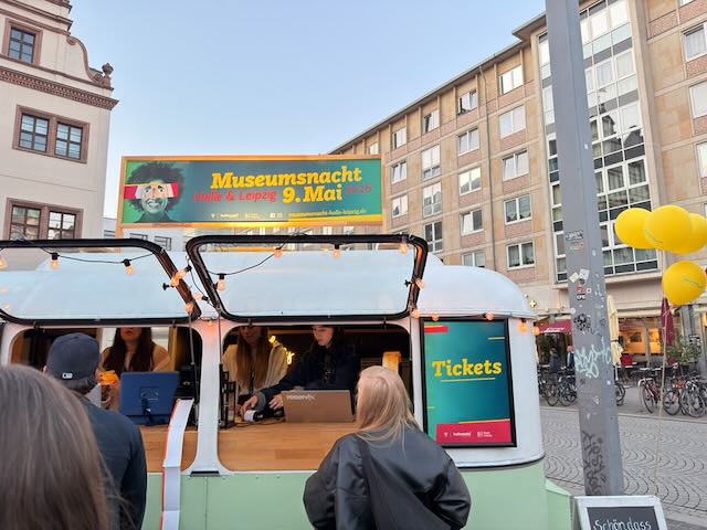 Menschen stehen an einem kleinen Anhänger an, aus dem heraus Tickets verkauft werden. Ein Schild auf dem Dach: "Museumsnacht Halle & Leipzig 9. Mai 2026"