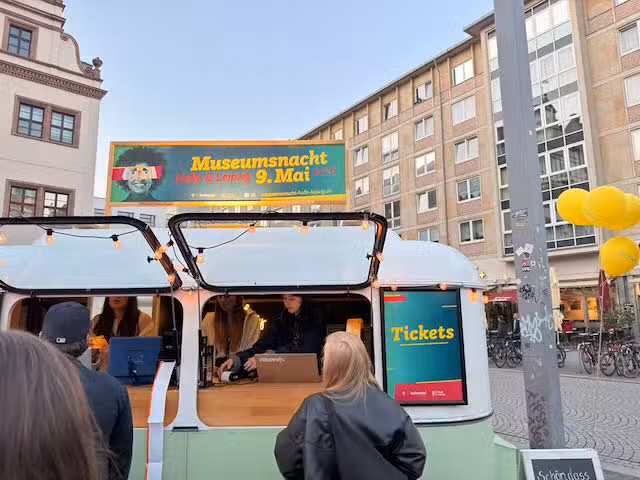 Menschen stehen an einem kleinen Anhänger an, aus dem heraus Tickets verkauft werden. Ein Schild auf dem Dach: "Museumsnacht Halle & Leipzig 9. Mai 2026"