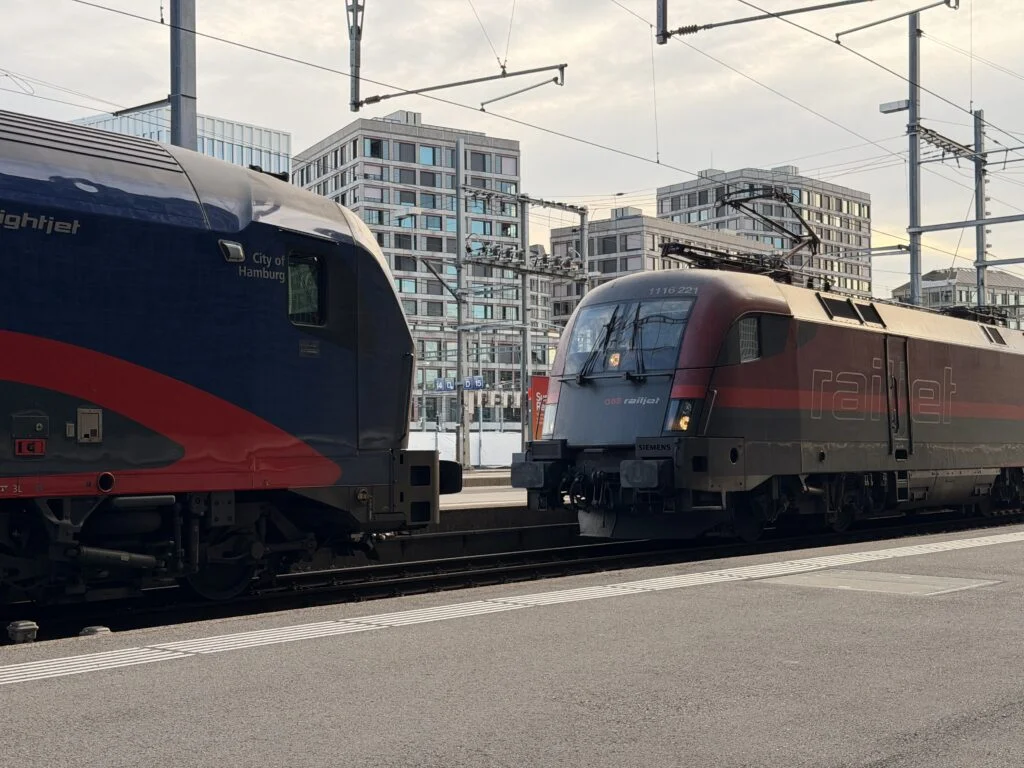 Zwei Züge der ÖBB Railjet stehen nebeneinander an einem Bahnsteig in Zürich, dahinter moderne Wohngebäude und Oberleitunsmasten.