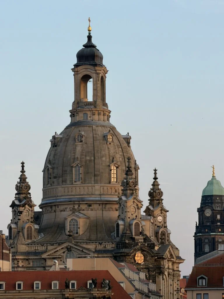 Barocke Kuppelkirche mit goldenem Kreuz und umliegenden Gebäuden in einer europäischen Stadt.