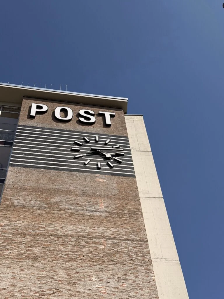 Fassade eines Postgebäudes mit großen weißen Buchstaben und einer Uhr unter klarem blauem Himmel.