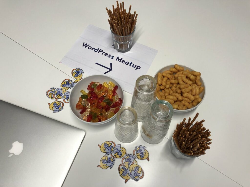 An assortment of snacks, some glasses and Wapuu stickers next to a printed sign "WordPress Meetup"