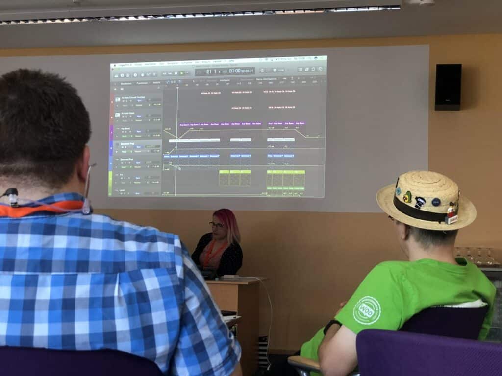 Laura, a women with pink hair, sits below a projector screen, showing the interface of Logic Pro