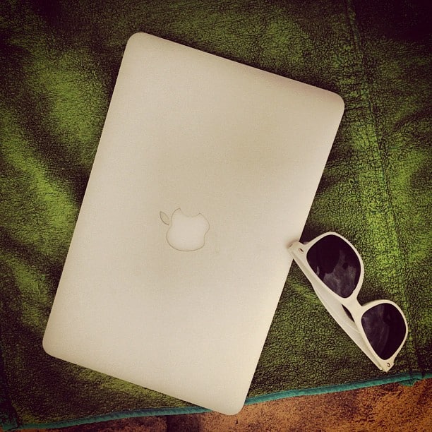 A silver MacBook and white sunglasses on a green towel. Next to the towel, some sand peaks out