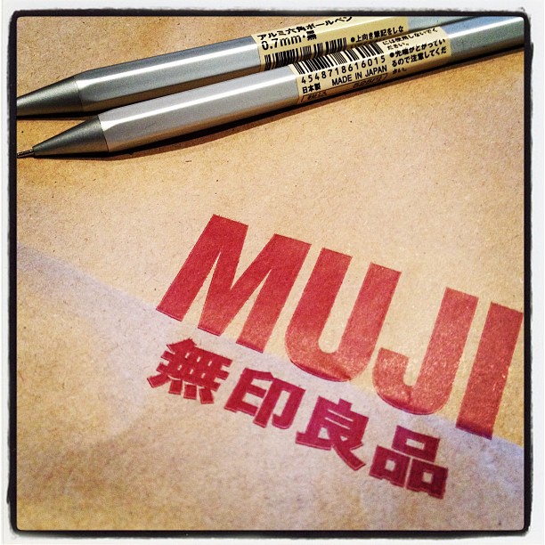 Two aluminium pens on a brown paper bag with a bright red "Muji" logo printed on the bag