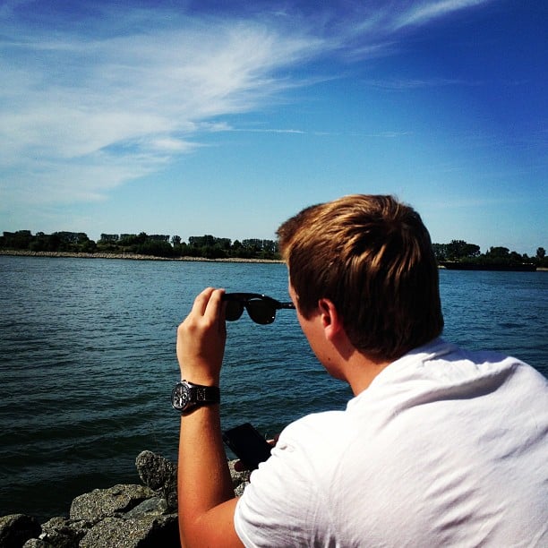 A man sitting next to a river, looking at his sunglasses in his hands