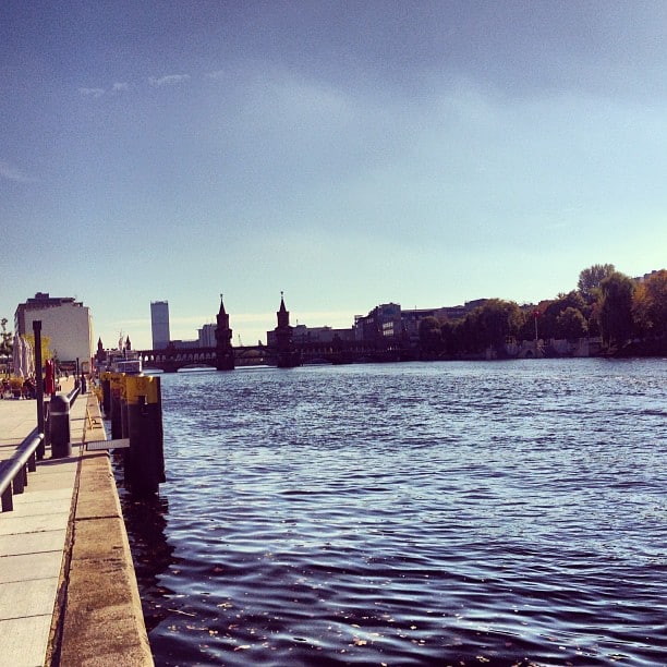 A path along a body of water, a red brick bridge in the background