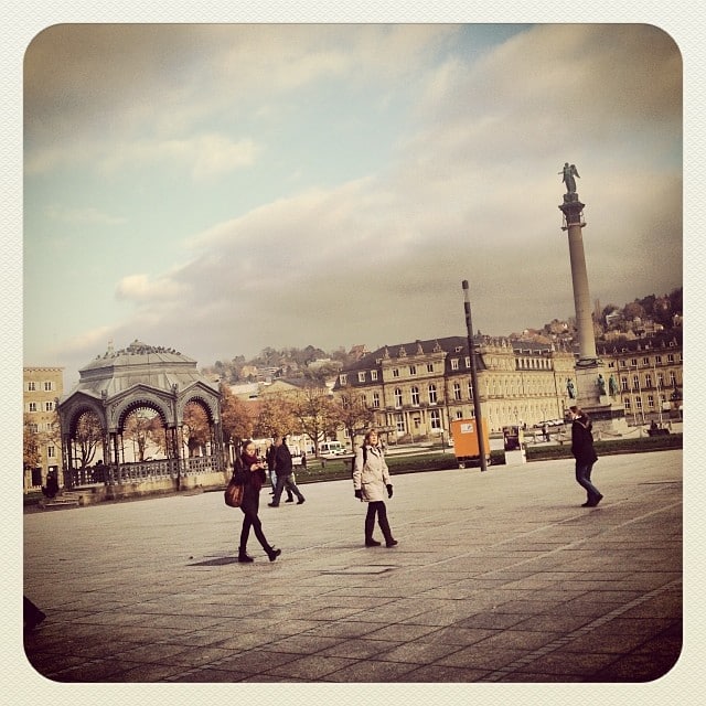 pedestrians in the foreground, a pavilion in the back, together with a tall column/memorial and some old buildings