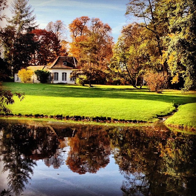 Scenic view of a park in fall. Trees in bright red and orange. A small pond in the foreground, followed by a green field and a small house