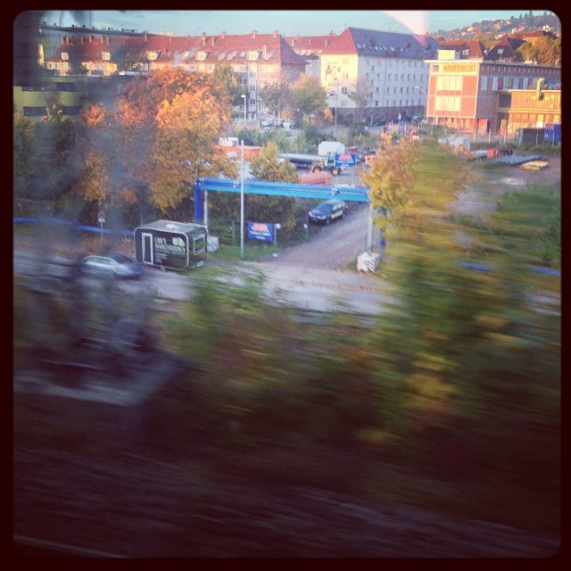 Photo taken from a moving train. Some bushes next to the train track in the foreground, building in the back