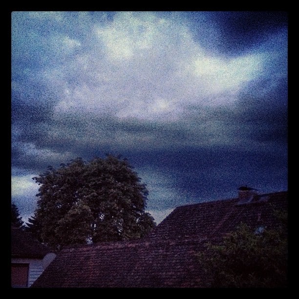 View out a window looking over some roofs and a big tree, very dark clouds above