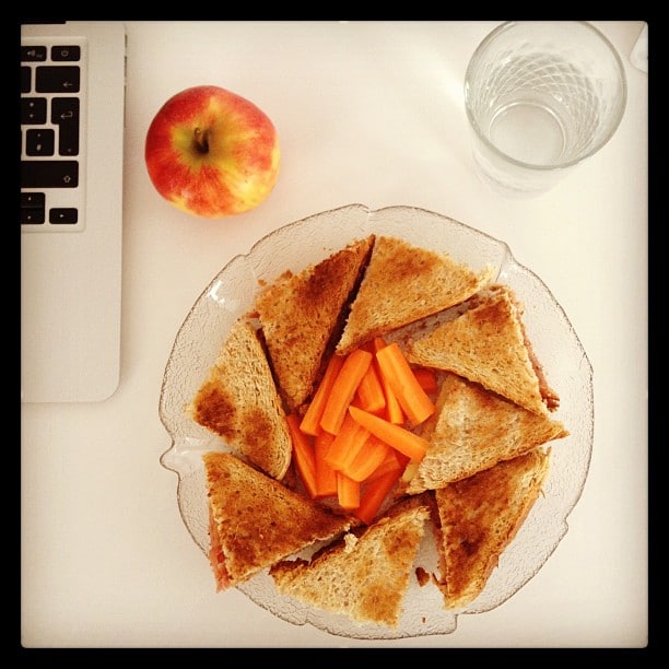 Top view of a white desk with a MacBook in the top left corner, next to an apple, a glas of water, and a plate of sandwiches with carrot sticks