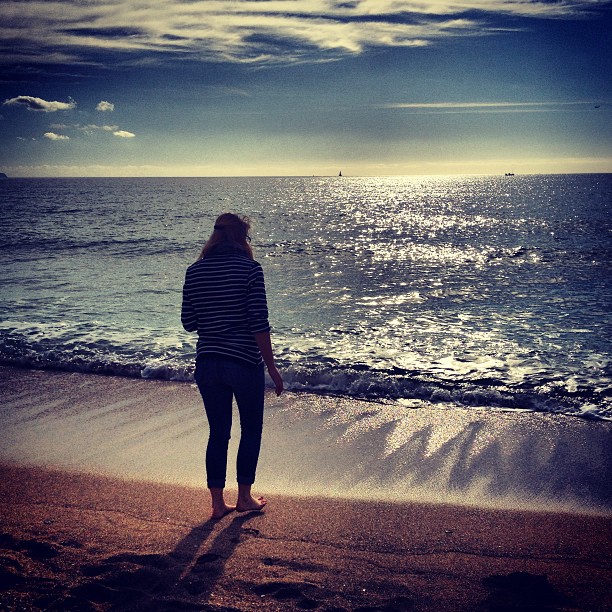 A woman, barefoot in street clothes walking on the beach toward the sea