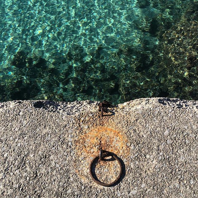 Top view of a harbour wall in the lower half of the image with clear water in the upper half. Centered on the wall a ring to tie down boats