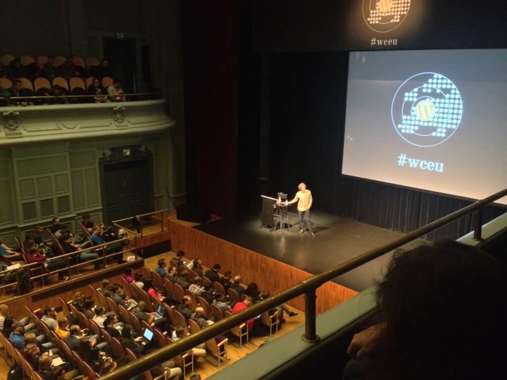 A man on stage, rows of chairs with people in front of him an on balconies on both sites. The projector screen behind him reads "#wceu"